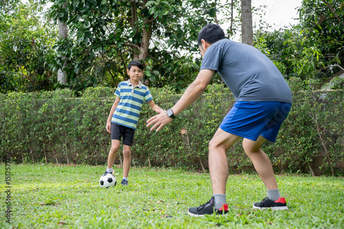 Father and son playing soccer or football together in their home's backyard or green grass field.
