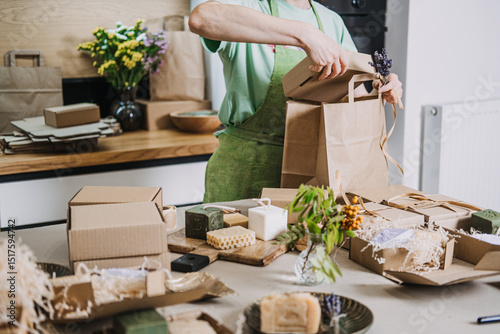 Gift boxes with handmade soaps are prepared on a kitchen table while a person ties a kraft bag. DIY business visuals, handmade product packaging, entrepreneur life, craft fair prep