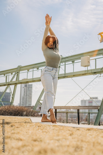Dancer stands barefoot in grass with arms raised under bright sky