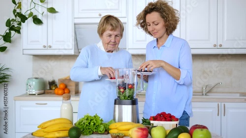 Elderly mother and adult daughter adding blueberries to blender, preparing healthy smoothie with various fruits and vegetables in white kitchen