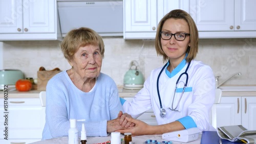 Caring female physician gently holding hands of senior woman, providing compassionate medical care and emotional support within cozy home kitchen environment