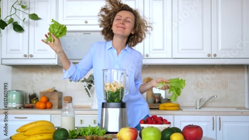 Cheerful woman dances with lettuce leaves in her hands, preparing a healthy green smoothie in her modern kitchen, surrounded by fresh fruits