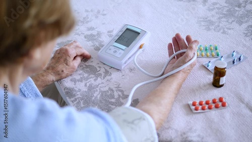 Senior woman using a digital blood pressure monitor at home, with medication blister packs and pill bottles visible on the table beside her