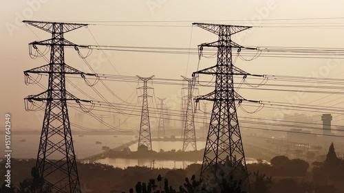 Power transmission towers and high-voltage lines under sunrise silhouette, dramatic long-exposure photography with warm golden tones, aerial view of energy infrastructure in natural landscape