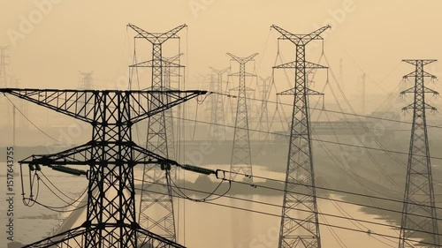 Power transmission towers and high-voltage lines under sunrise silhouette, dramatic long-exposure photography with warm golden tones, aerial view of energy infrastructure in natural landscape