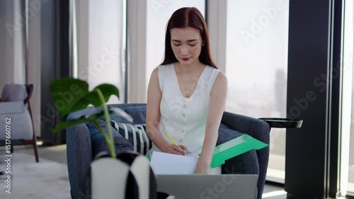 An adult asian woman engrossed in her work is checking data on her laptop to prepare a detailed report for her superior in a chic, contemporary office while enjoying natural light from large windows
