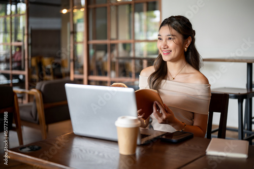 Asian woman looking away from book while sitting in front of a laptop with coffee cup aside in cafe.