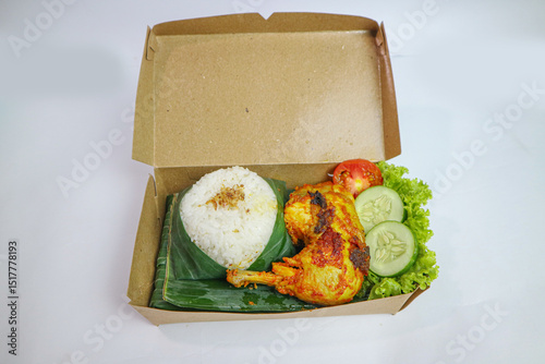 Nasi Kotak featuring ayam bakar (roasted chicken), served with a mound of white rice and fresh vegetables like cucumber and tomato. flat lay photo concept