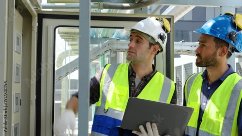 Two engineers in safety uniform checking building exterior power system at construction site rooftop, laptop in hand, sharing expertise in a moment of industrial collaboration, Worker maintenance