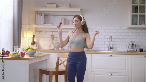 Young Asian athletic woman wearing headset sings while listening to music and cooking healthy fruit salad and protein shake in home kitchen. Active lifestyle, wellness, diet, and nutrition concept.