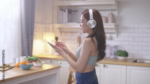 Young Asian athletic woman wearing headset sings while listening to music and cooking healthy fruit salad and protein shake in home kitchen. Active lifestyle, wellness, diet, and nutrition concept.