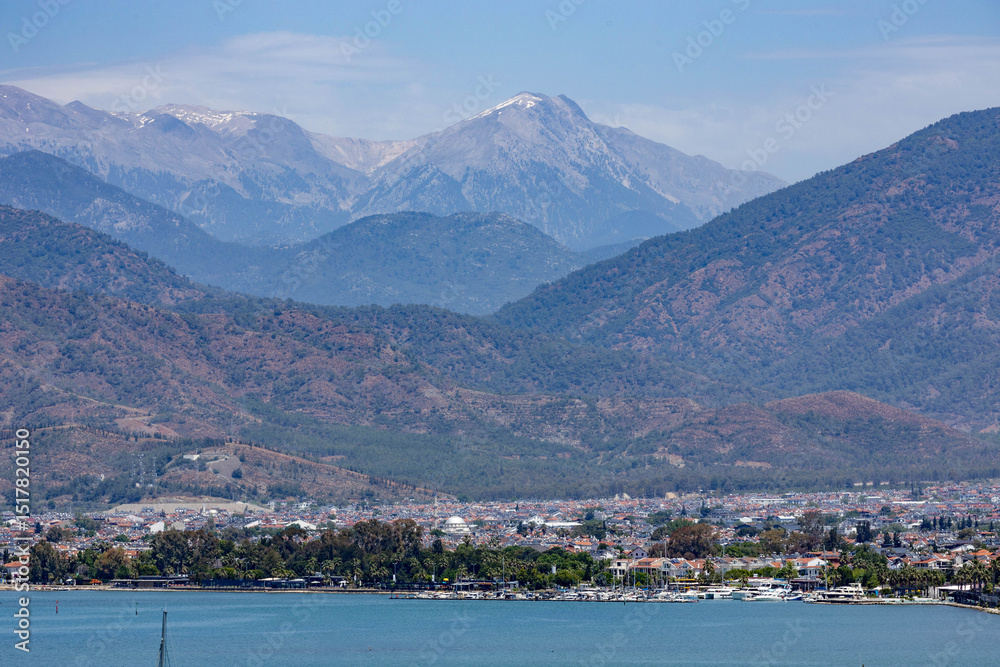 Fototapeta premium Panoramic view of Fethiye with waterfront, cityscape, and snow-capped Taurus Mountains under blue sky. Muğla, Turkey, Mediterranean region. Sunny spring day.
