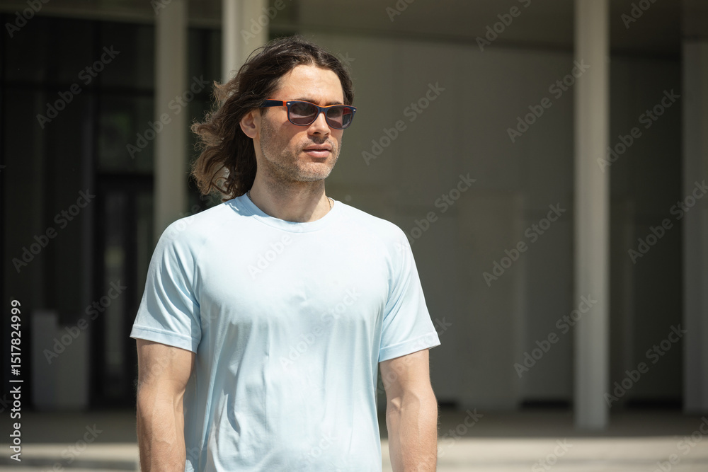 Athletic caucasian man with long curly hair wearing sunglasses and light blue T-shirt walks confidently outdoors. His thoughtful expression highlights the calm and relaxed atmosphere of sunny day Athletic caucasian man with long curly hair wearing sunglasses and light blue T-shirt walks confidently outdoors. His thoughtful expression highlights the calm and relaxed atmosphere of sunny day