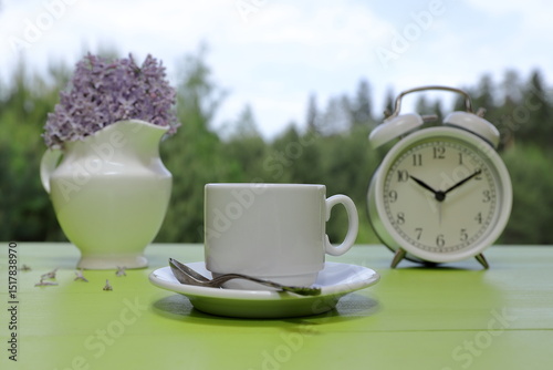 A white cup of coffee stands next to a retro alarm clock and a white jug of lilac flowers on a green wooden desk against a summer woodland background. 