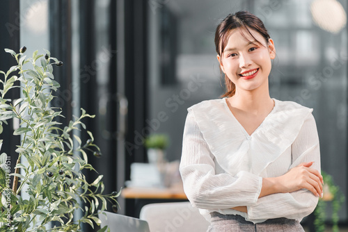 Confident young Asian woman in white blouse smiling with arms crossed, sitting in modern office, expressing positivity and professionalism