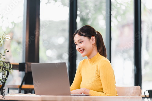 Free Download Young Asian woman in yellow sweater smiling while working on laptop at modern bright office, natural light, casual workspace, positive mood, productivity, technology - GFXLoad