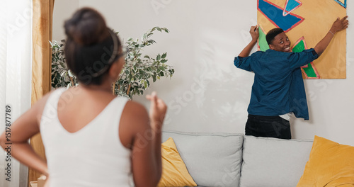 A couple in love decorates their first apartment. The man hangs a painting on the wall while the woman gestures how to adjust it. Around them are unpacked boxes after the move.