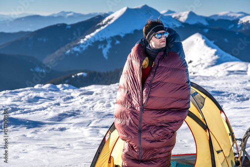 Man standing outside tent in snowy mountains, wrapped in sleeping bag, warming up in cold morning light