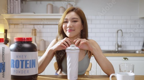 A young, athletic Asian woman in stylish sportswear prepares a nutritious protein shake in her modern kitchen while live-streaming a health-focused video tutorial. With a cheerful smile