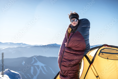 Man standing outside tent in snowy mountains, wrapped in sleeping bag, warming up in cold morning light