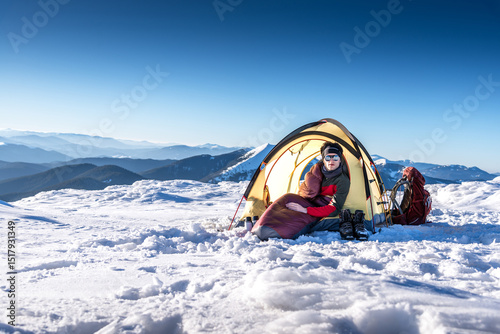 Male backpacker relaxing in a sleeping bag inside a tent on a snowy alpine ridge at sunrise. Winter camping, solo trekking, mountaineering, and cold weather outdoor adventure concept.