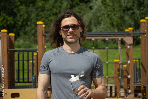 Portrait of happy caucasian man in casual attire stands in sunglasses at park holding water bottle, enjoying sunny day near kids playground equipment. Relaxed and contentment. Outdoors.