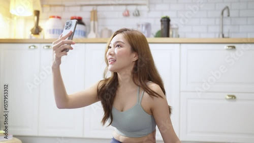 A young, healthy Asian woman in activewear takes a cheerful selfie or video call on her smartphone while relaxing after a yoga workout with her community in a cozy apartment, embracing wellness