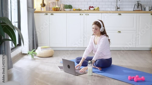 young Asian athletic woman wearing a sporty top and leggings practices yoga in her cozy living room while attentively listening to spiritual wellness lessons on her laptop. Focused on mindfulness