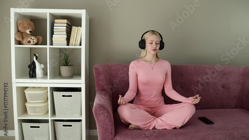 Blonde woman in pink loungewear sitting cross-legged on a purple couch, eyes closed and wearing headphones, meditating in a calm home environment with phone placed nearby