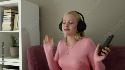 Close-up of a smiling blonde woman in pink loungewear sitting on a sofa, wearing black headphones and holding a smartphone while dancing to music