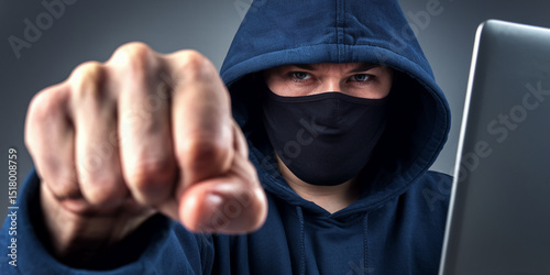 Close-up of fist and masked person in hoodie, laptop in background, symbolizing online threat, aggression, or cybercrime