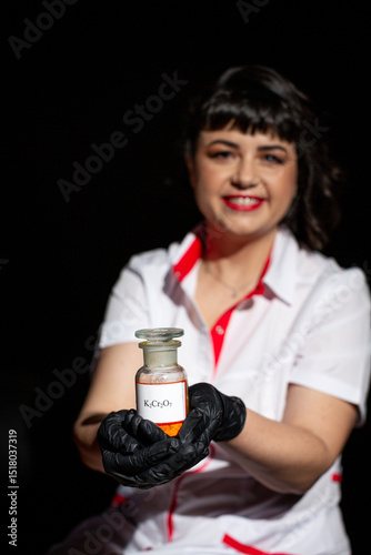 Chemistry teacher in white coat and black gloves holding glass jar with orange potassium dichromate powder on black background