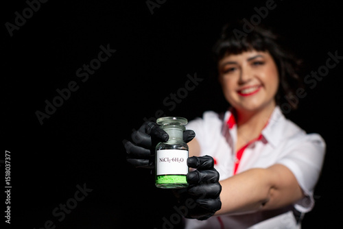 Chemistry teacher in white coat and black gloves holding glass jar with green powder of hexahydrate chloride on black background