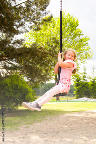 Smiling girl in pink outfit swinging on a rope swing at a sunny playground. Green trees and sand in background. Joyful childhood moment in nature.

