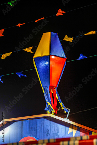 Large colorful balloon lantern in red, blue, and yellow hanging above wooden festival booth during nighttime Brazilian festa junina celebration, with decorative bunting flags strung overhead.