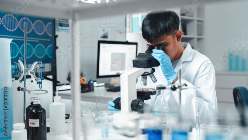 Focused Asian male scientist using microscope in modern laboratory with DNA structure chart in background. Concept of biotechnology, genetics research, molecular biology and advanced healthcare.