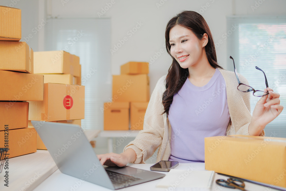 Young entrepreneur working in her home office, surrounded by cardboard boxes, using laptop and holding eyeglasses, managing her online business Young entrepreneur working in her home office, surrounded by cardboard boxes, using laptop and holding eyeglasses, managing her online business