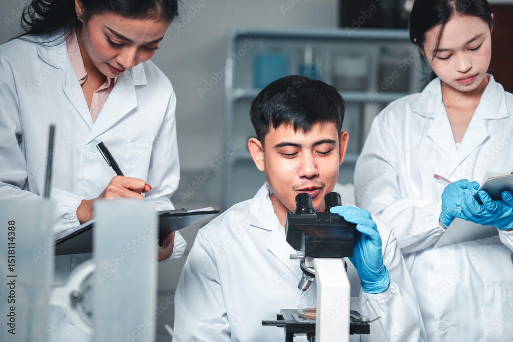 Three young scientists in lab coats collaborate on a research project using a microscope and taking notes, surrounded by lab equipment and blue liquids, showing teamwork and scientific analysis.