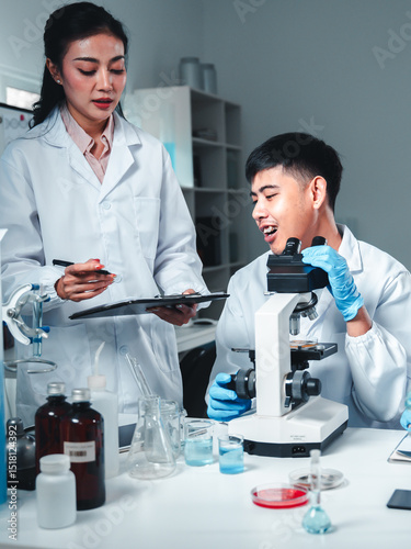 Group of young scientists in lab coats collaborate in a high-tech laboratory, analyzing samples through a microscope and documenting results, symbolizing innovation, teamwork, and modern research.