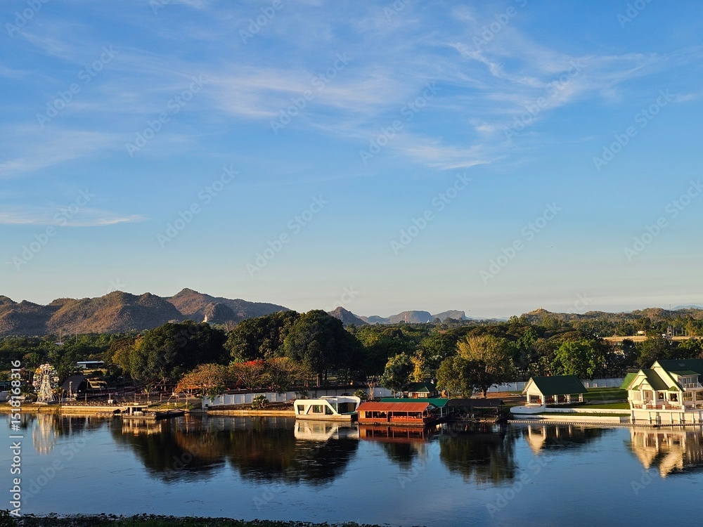 A peaceful riverside scene shows buildings and trees reflected in calm water, with mountains in the background and a clear blue sky above. A peaceful riverside scene shows buildings and trees reflected in calm water, with mountains in the background and a clear blue sky above.