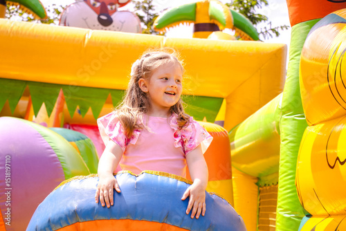 Happy little girl playing on a colorful inflatable bouncy castle. Child in pink outfit jumps with joy. Bright outdoor setting with soft light and vivid colors.


