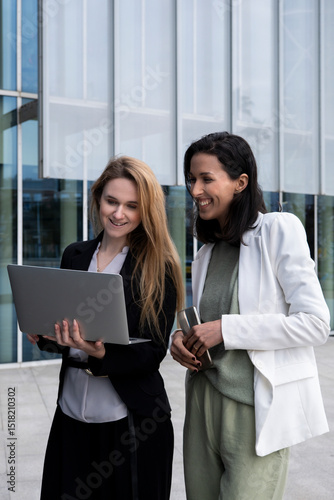 Two women in their 30s from different ethnicities collaborating outdoors on a laptop in a modern corporate setting
