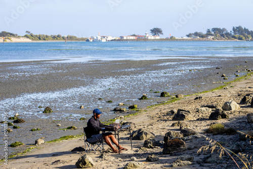 A man with a laptop working on a beach or bay at low tide.  Full-time RV nomad lifestyle, working in shorts from his home office.