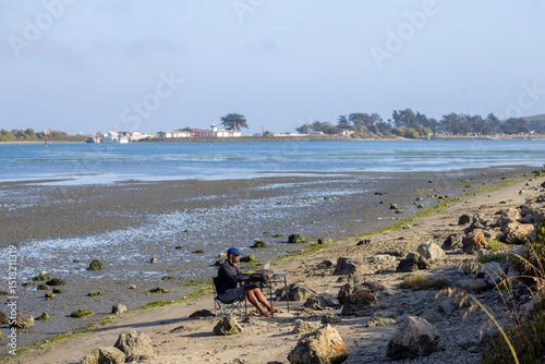 A man with a laptop working on a beach or bay at low tide.  Full-time RV nomad lifestyle, working in shorts from his home office.