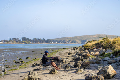 A man with a laptop working on a beach or bay at low tide.  Full-time RV nomad lifestyle, working in shorts from his home office.
