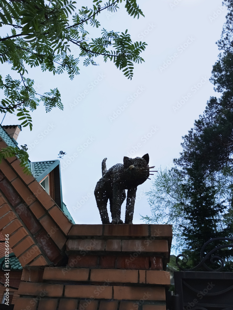 Fototapeta premium Metal statue figurine cat on the brick wall of the house. Summer twilight.