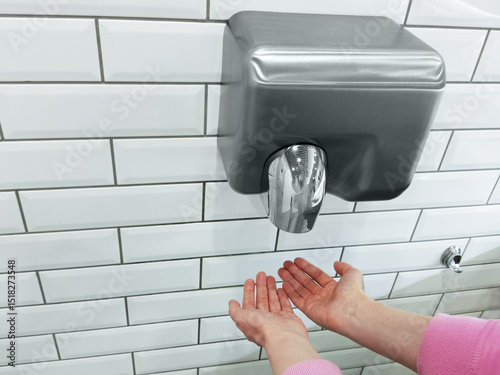 Caucasian woman's hands are dried by a wall-mounted dryer in the toilet, the wall is white tiled.