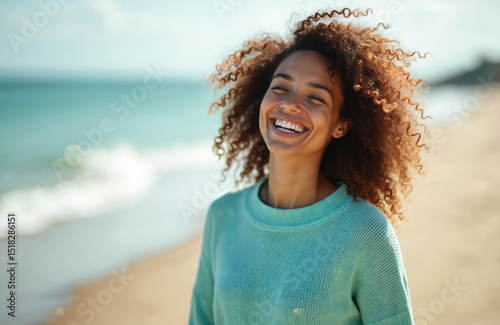 Happy woman enjoys sunny day at beach with ocean view. Portrait of smiling person with natural curly hair. Joyful adult female with white teeth smiles, embodies summer joy, happiness, wellbeing,