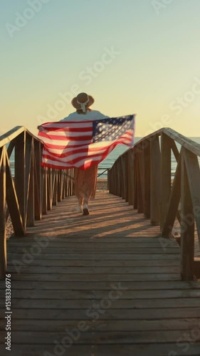 Female runner wearing wide-brimmed sunhat waving american flag while crossing wooden pier during golden sunset by coastline. Usa celebrate 4th of july. Independence day concept