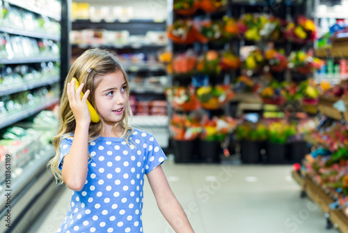 Female child holding banana like phone in supermarket produce aisle with flower displays